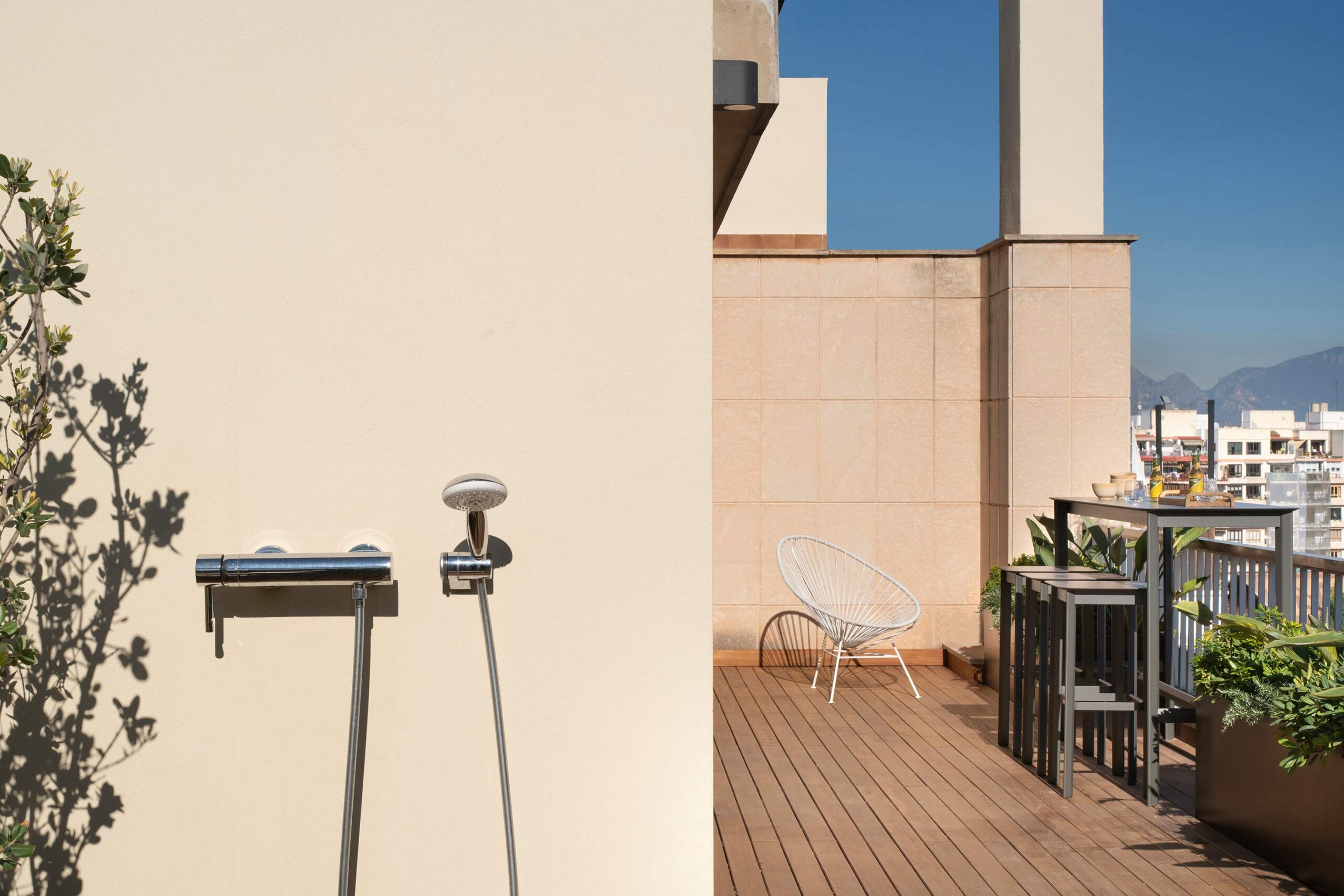 Terraza con ducha exterior en pared beige, silla blanca, barra alta y montañas al fondo