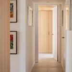 Pasillo blanco con cuadros de colores, suelo de baldosa clara y armarios de madera al fondo