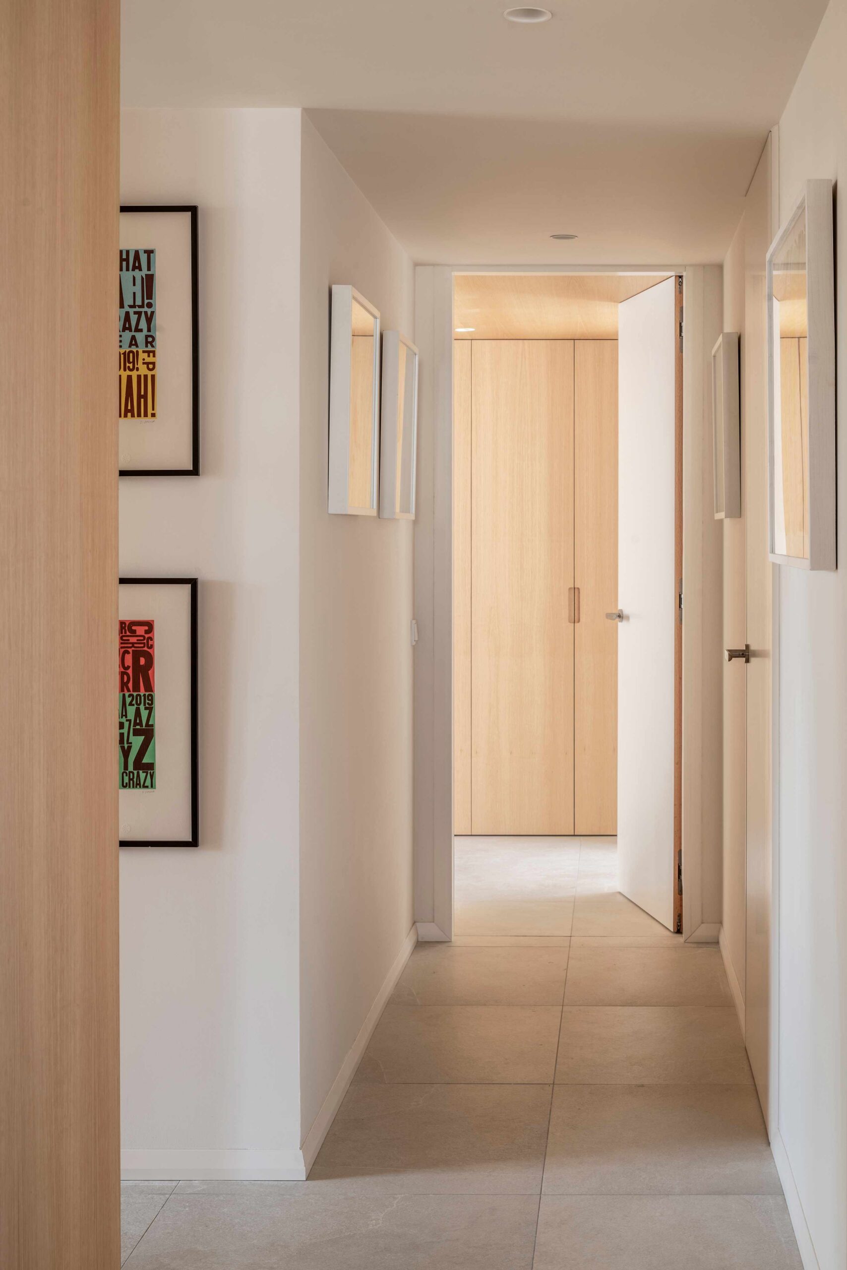 Pasillo blanco con cuadros de colores, suelo de baldosa clara y armarios de madera al fondo
