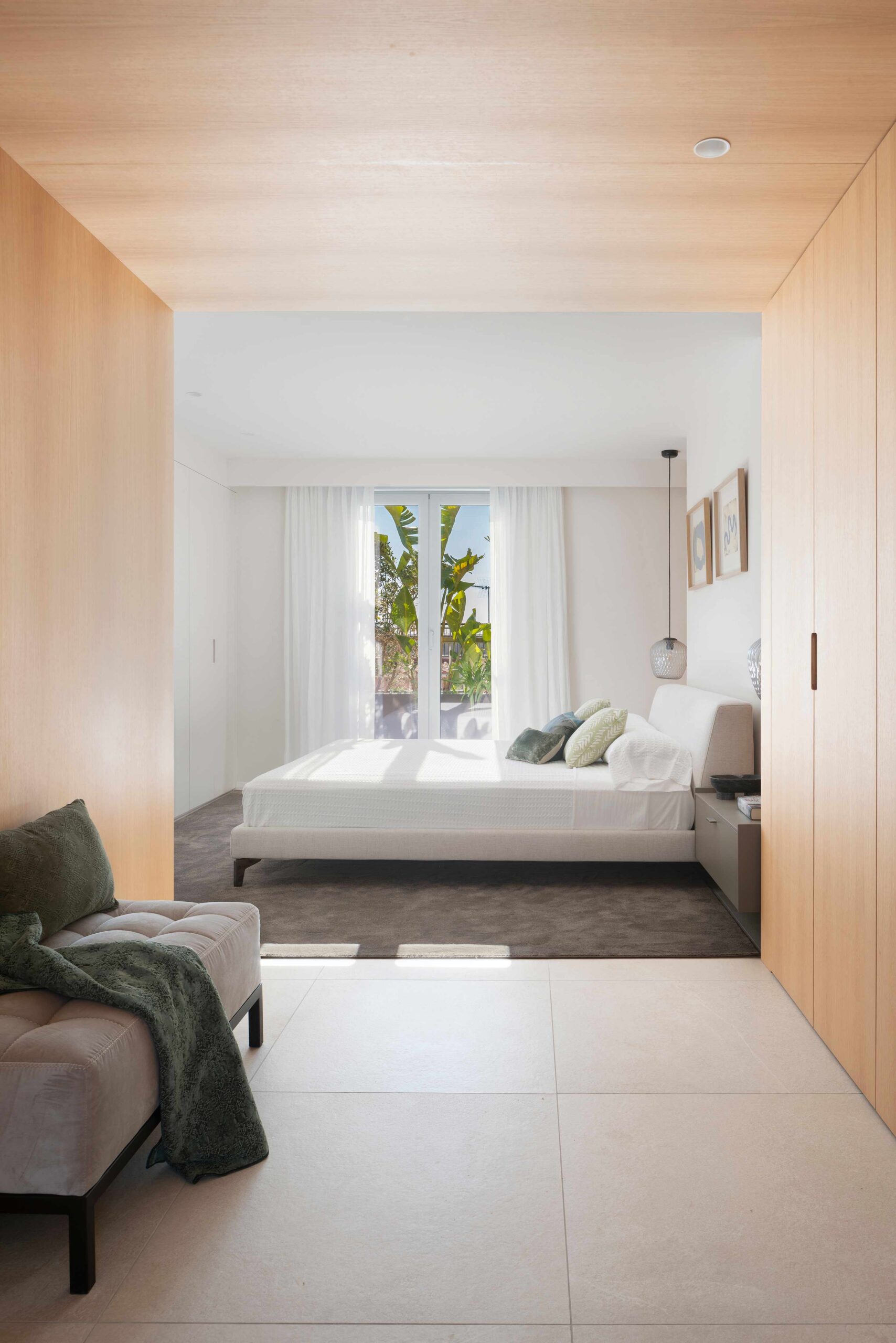 Dormitorio con cama blanca y ventanal a la terraza, visto desde un acceso con armarios de madera clara