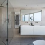 Baño con ducha acristalada, doble lavabo blanco suspendido, toallero radiador, bañera y ventana con cielo azul