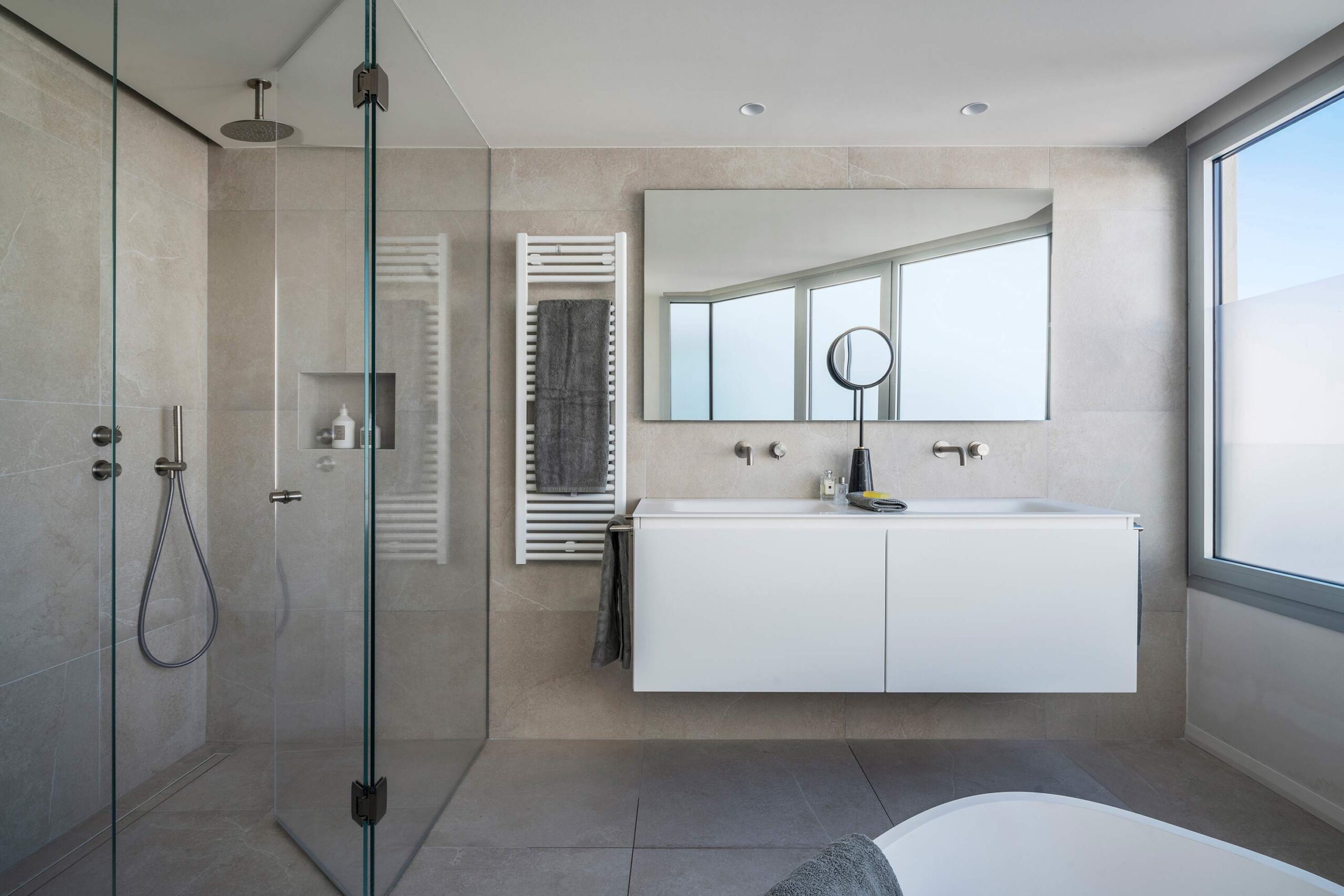 Baño con ducha acristalada, doble lavabo blanco suspendido, toallero radiador, bañera y ventana con cielo azul
