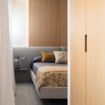Detalle de dormitorio con cabecero gris, panel de madera con luz indirecta, manta ocre y cortina blanca