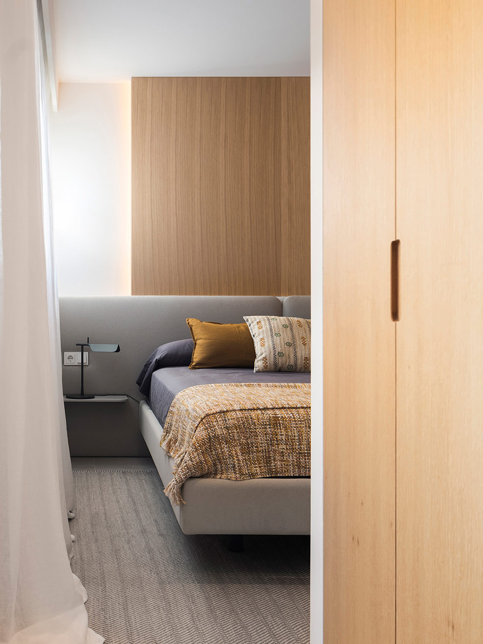 Detalle de dormitorio con cabecero gris, panel de madera con luz indirecta, manta ocre y cortina blanca