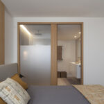 Dormitorio con cama de cabecero gris y paso de madera con vidrio esmerilado hacia el baño