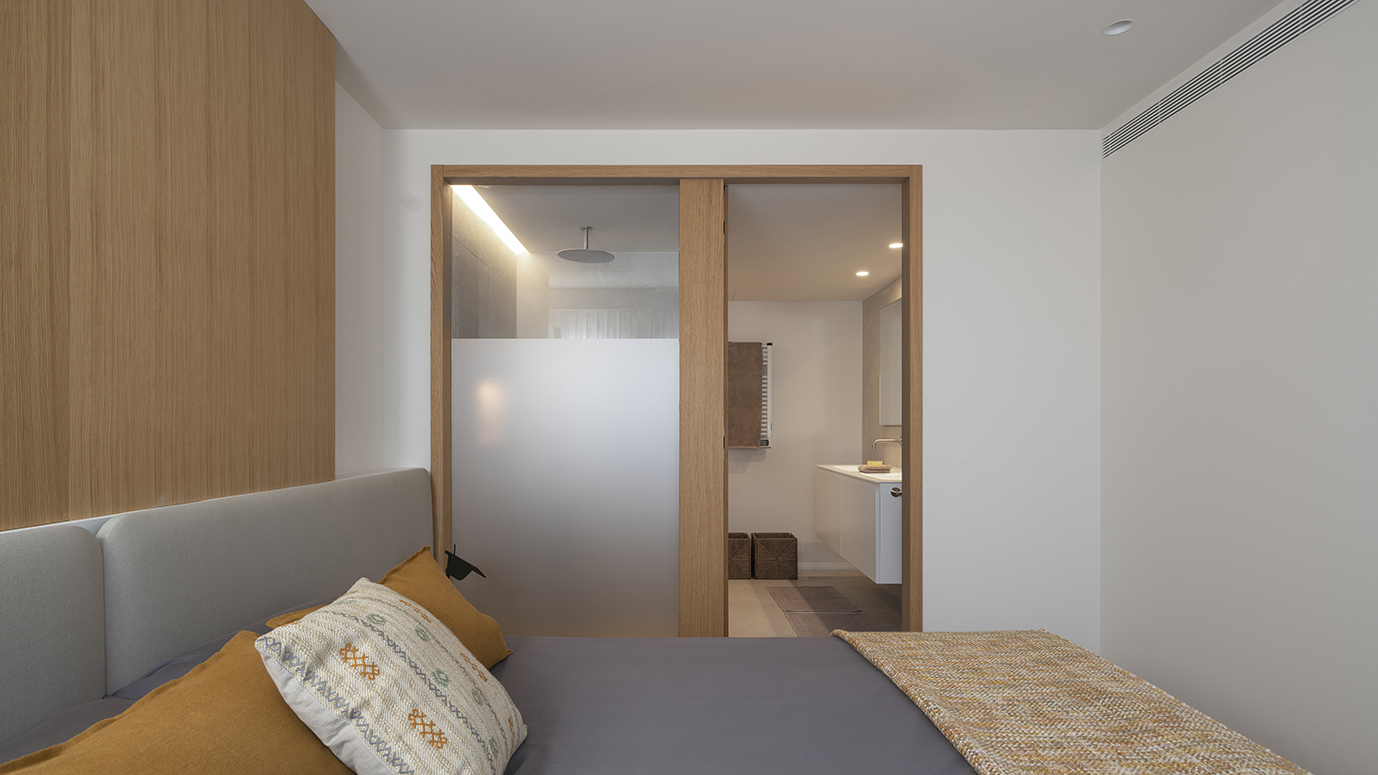 Dormitorio con cama de cabecero gris y paso de madera con vidrio esmerilado hacia el baño