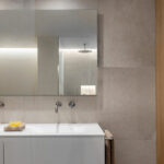 Baño con doble lavabo blanco, grifería empotrada en pared, gran espejo y revestimiento gris de aspecto pétreo