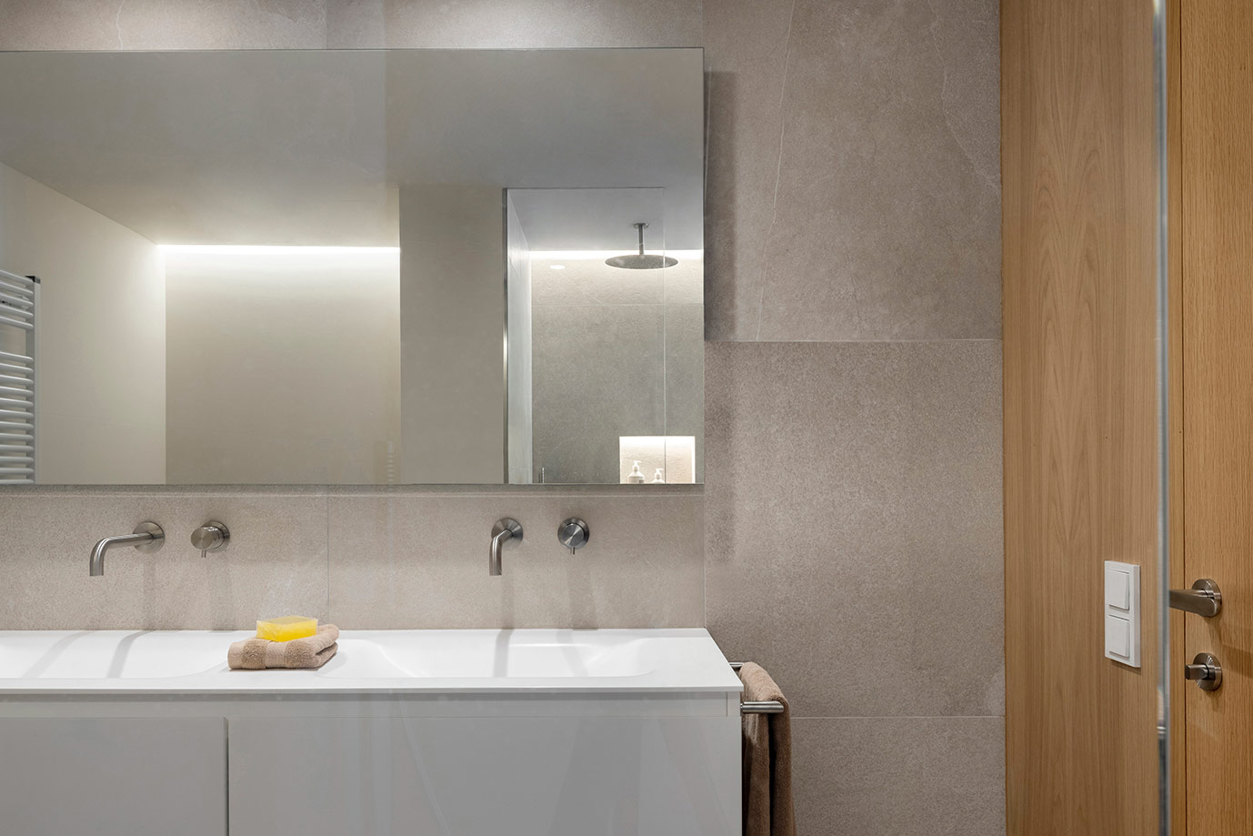 Baño con doble lavabo blanco, grifería empotrada en pared, gran espejo y revestimiento gris de aspecto pétreo