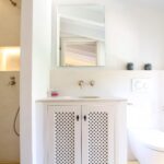 Baño con lavabo sobre mueble blanco de celosía, grifería color cobre, ducha de microcemento y espejo rectangular