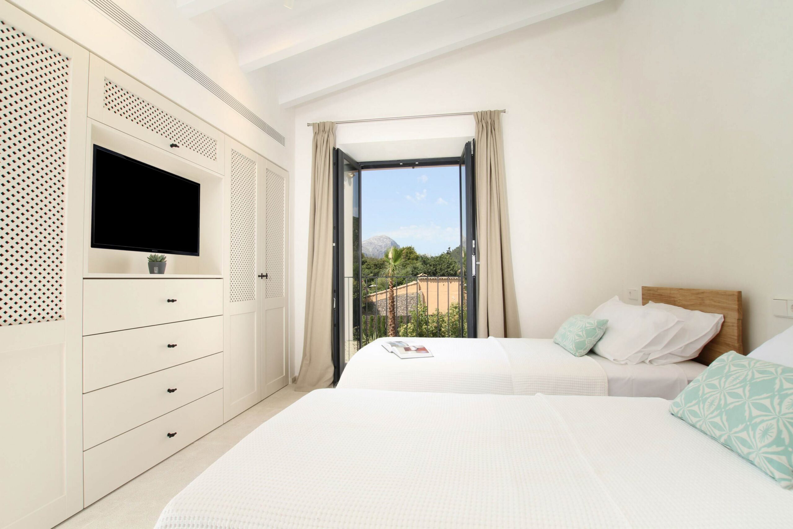 Dormitorio de dos camas con balconera abierta a la montaña, armario blanco y cojines verde agua