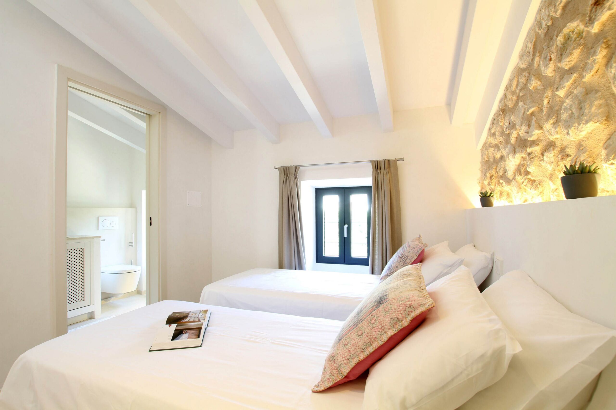 Habitación con dos camas bajo vigas blancas, pared de piedra vista iluminada y baño en suite