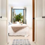 Puertas de celosía blancas enmarcan un dormitorio con cama blanca y balconera abierta a plantas tropicales