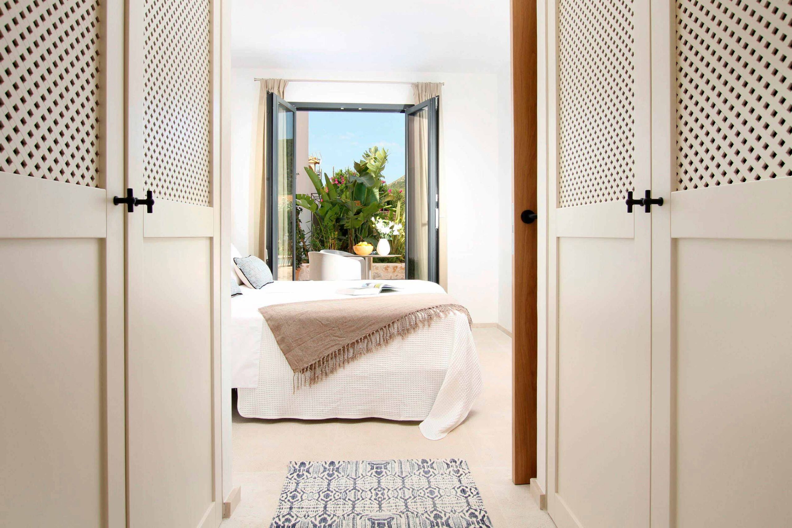Puertas de celosía blancas enmarcan un dormitorio con cama blanca y balconera abierta a plantas tropicales