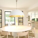 Comedor con mesa redonda y sillas de madera clara bajo lámpara blanca, cocina abierta al fondo