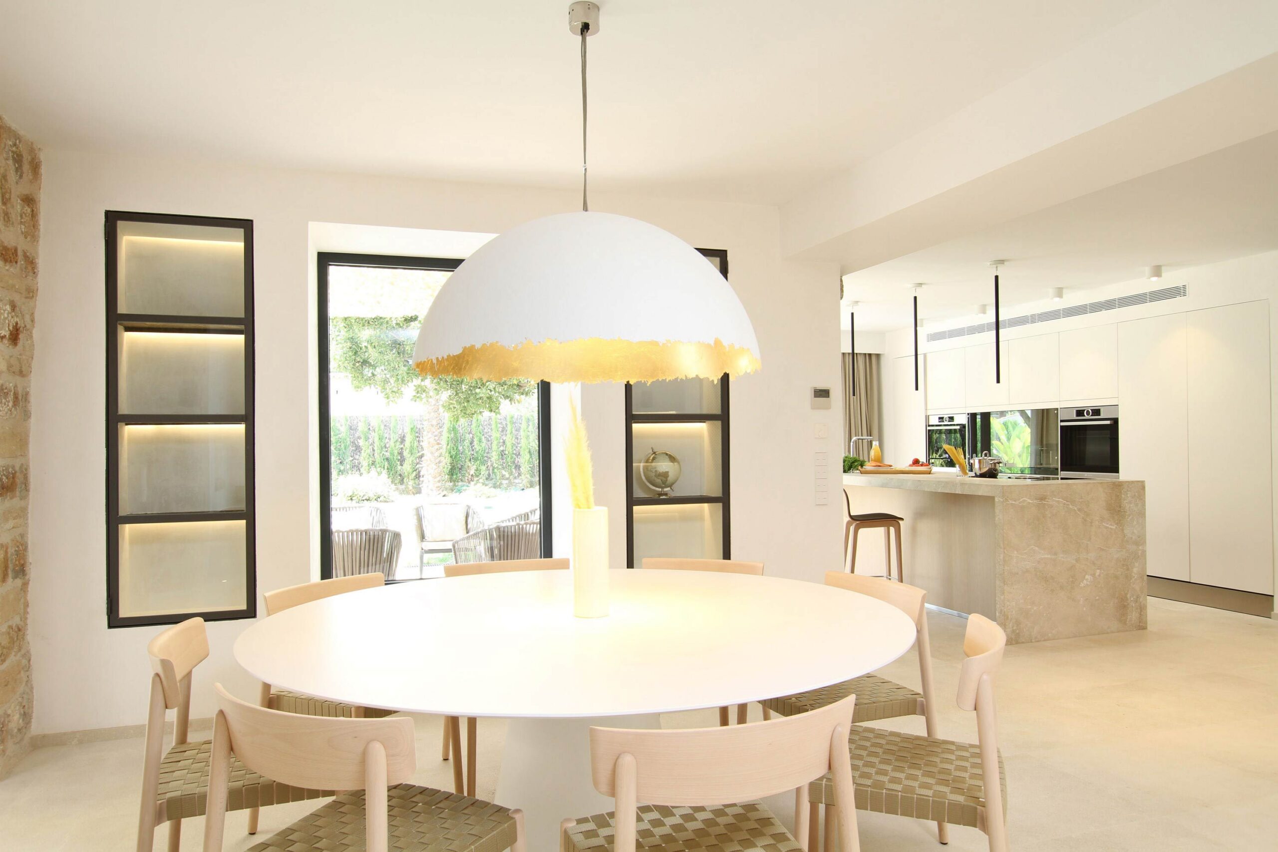 Comedor con mesa redonda y sillas de madera clara bajo lámpara blanca, cocina abierta al fondo