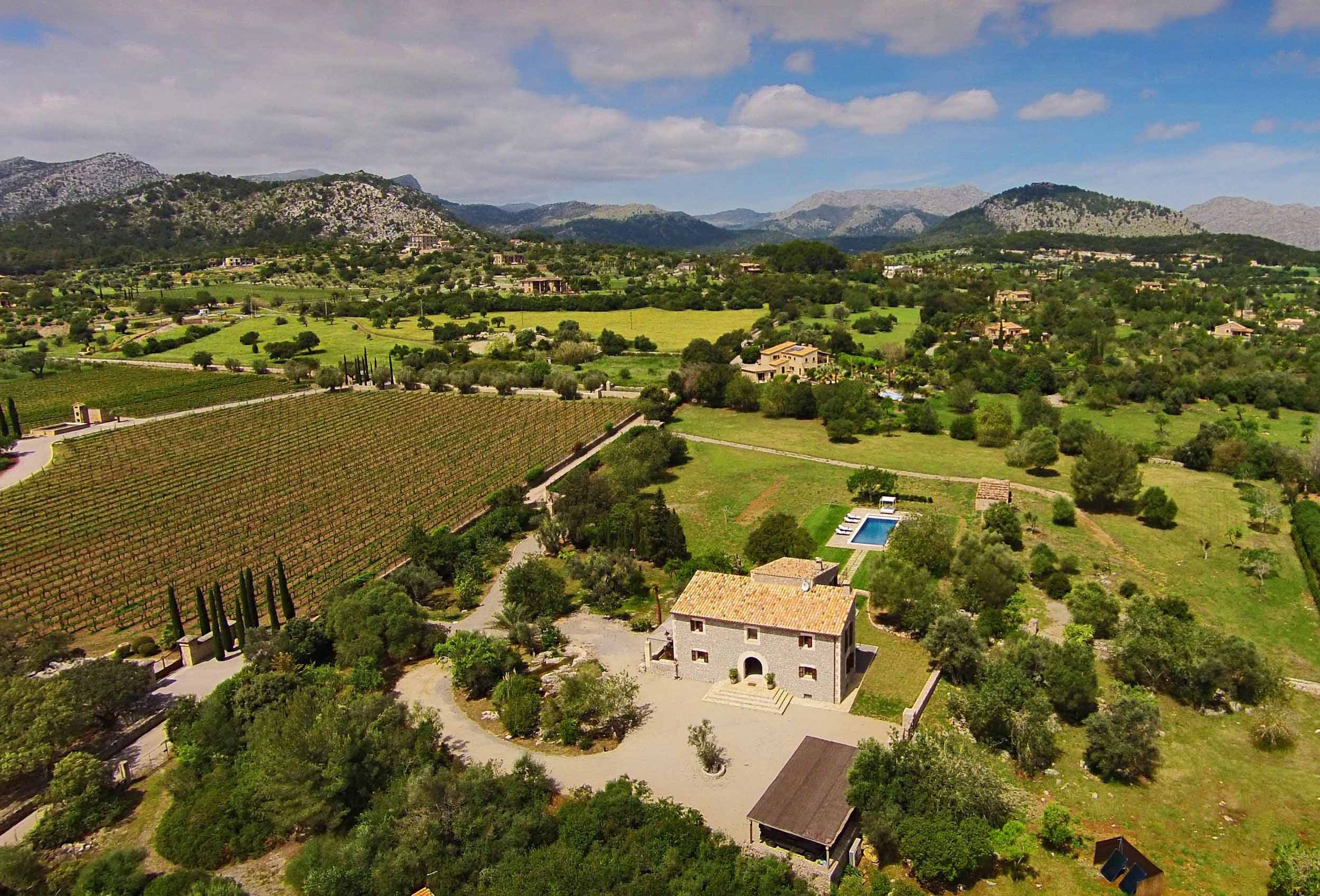 Vista aérea de casa de piedra con tejado de teja, jardines, piscina, viñedo y montañas al fondo