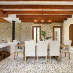 Comedor con mesa de madera, sillas blancas, pared de piedra, vigas vistas y suelo de guijarros