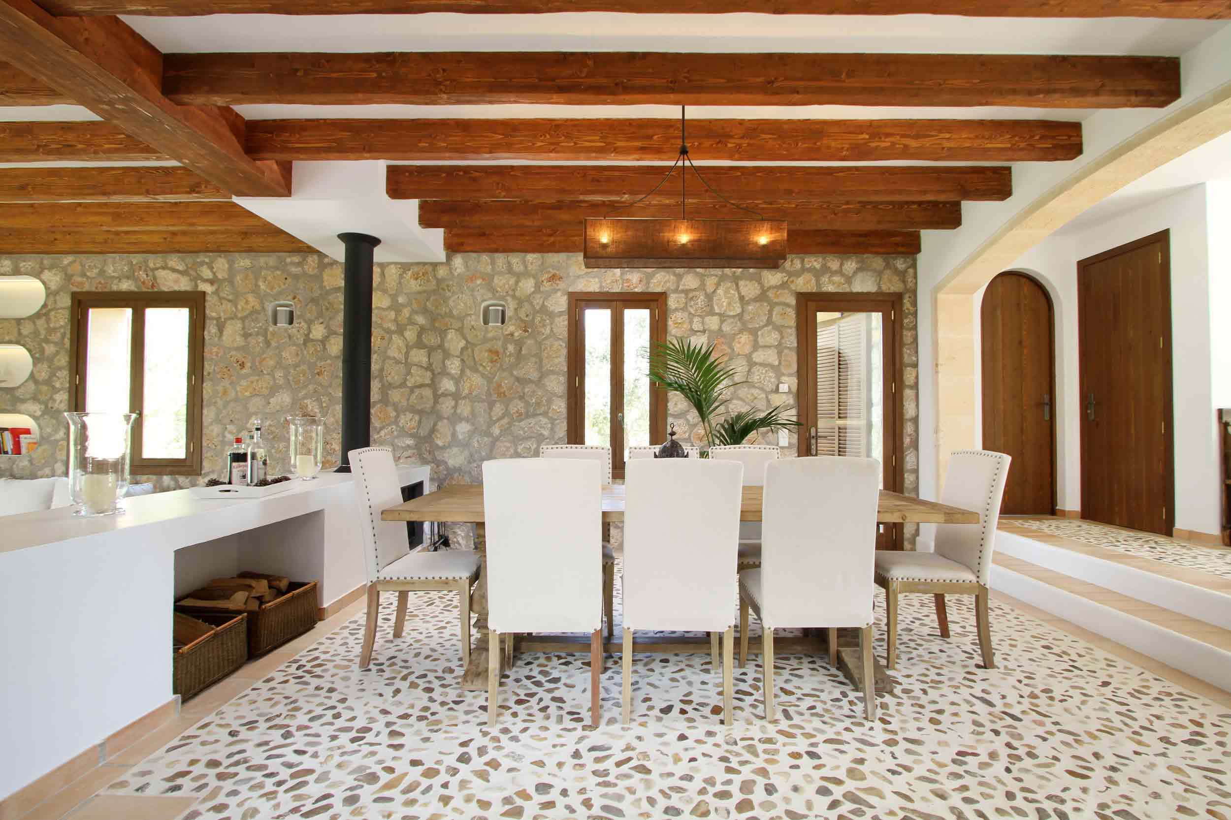 Comedor con mesa de madera, sillas blancas, pared de piedra, vigas vistas y suelo de guijarros
