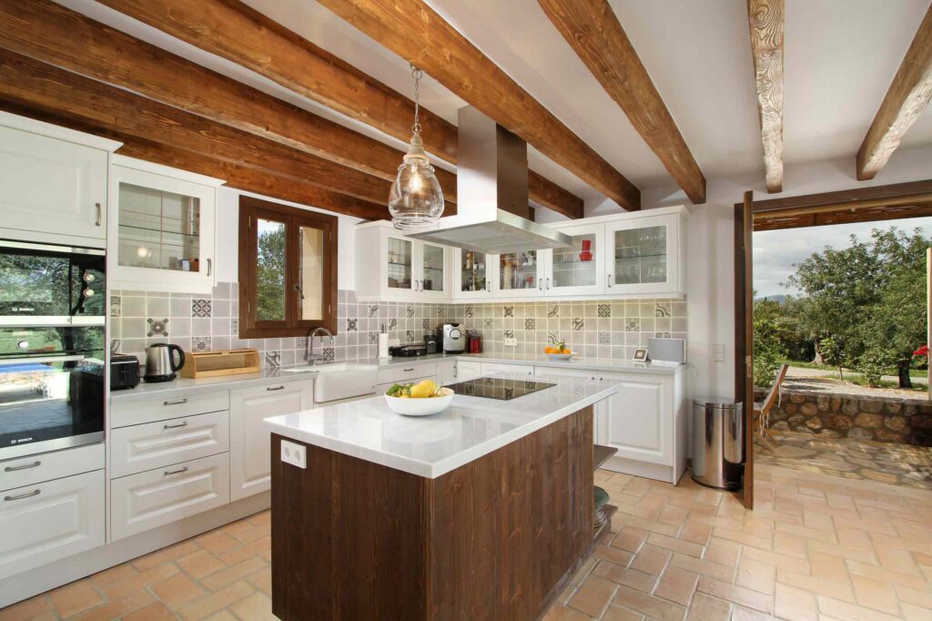Cocina con isla de madera oscura y encimera blanca, armarios blancos, vigas vistas y suelo de terracota