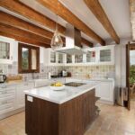 Cocina con isla de madera oscura y encimera blanca, armarios blancos, vigas vistas y suelo de terracota