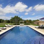 Piscina alargada de mosaico azul con tumbonas, cama con dosel y caseta de piedra entre prados verdes