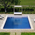 Piscina rectangular vista desde arriba con borde claro, pavimento de piedra irregular, tumbonas y cama con dosel