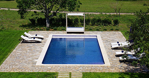 Piscina rectangular vista desde arriba con borde claro, pavimento de piedra irregular, tumbonas y cama con dosel