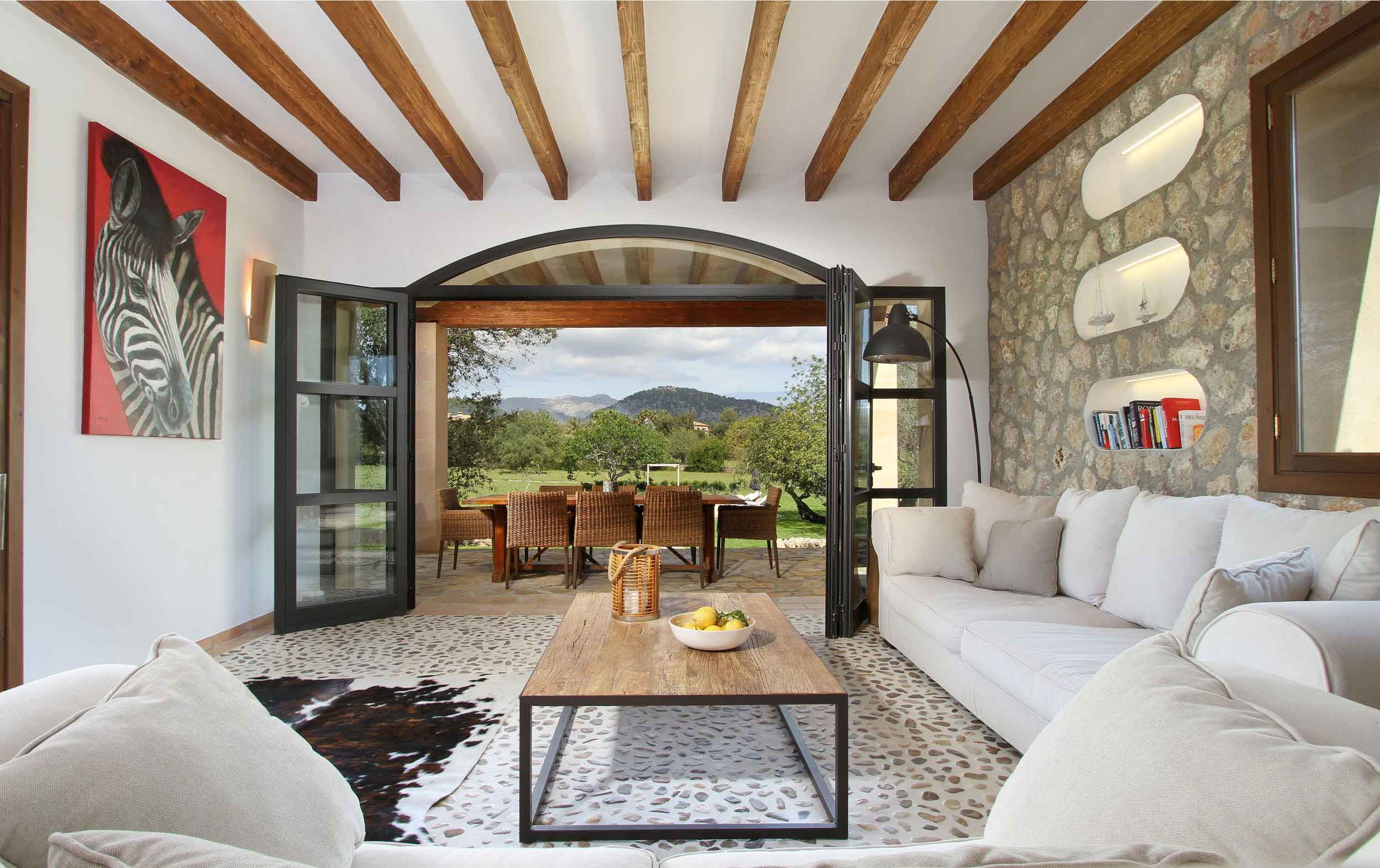 Salón con sofás blancos, pared de piedra, vigas de madera y terraza con vistas a la montaña