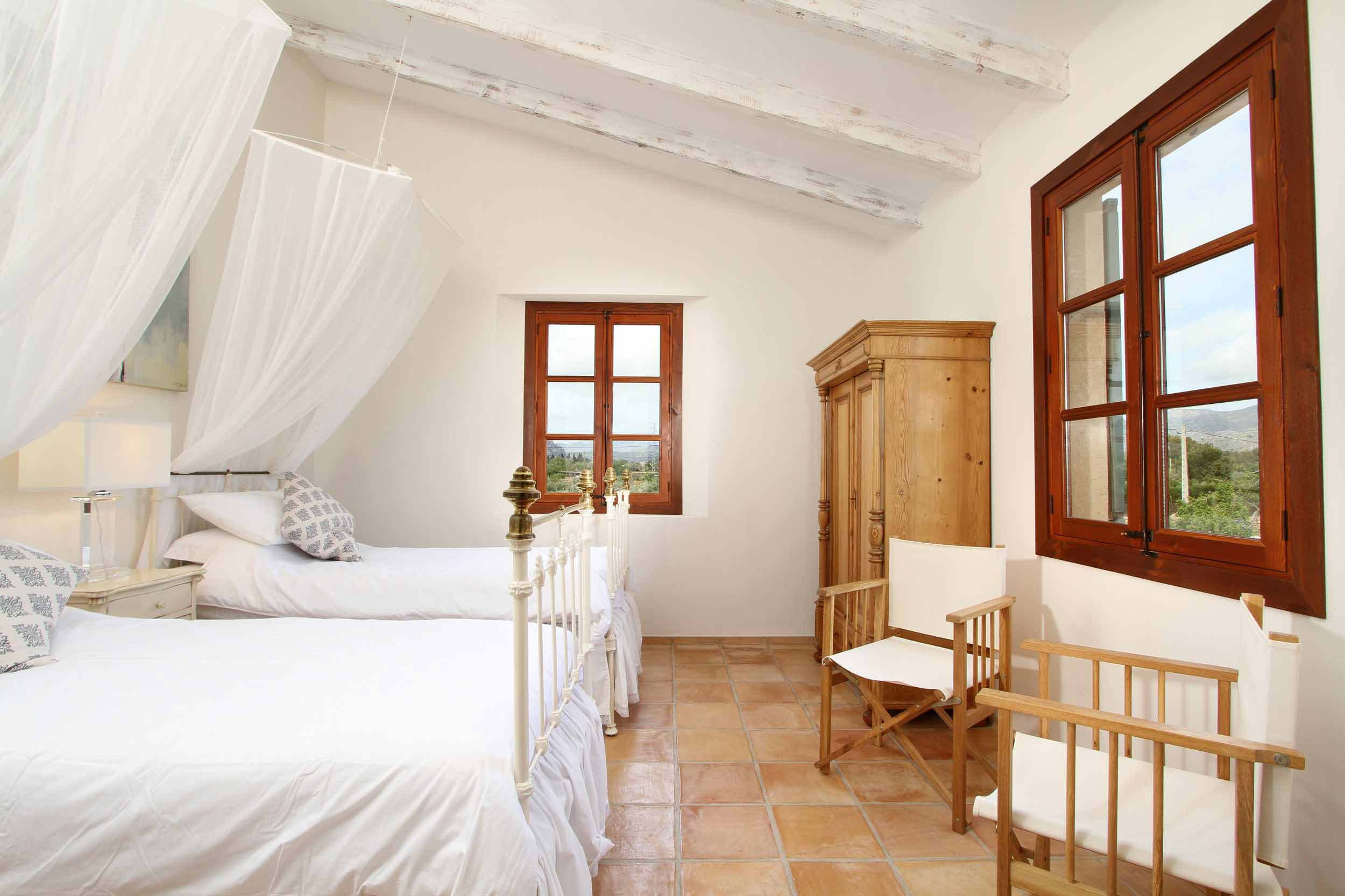 Dormitorio con dos camas de hierro, doseles blancos, ventanas de madera con vistas y suelo de terracota