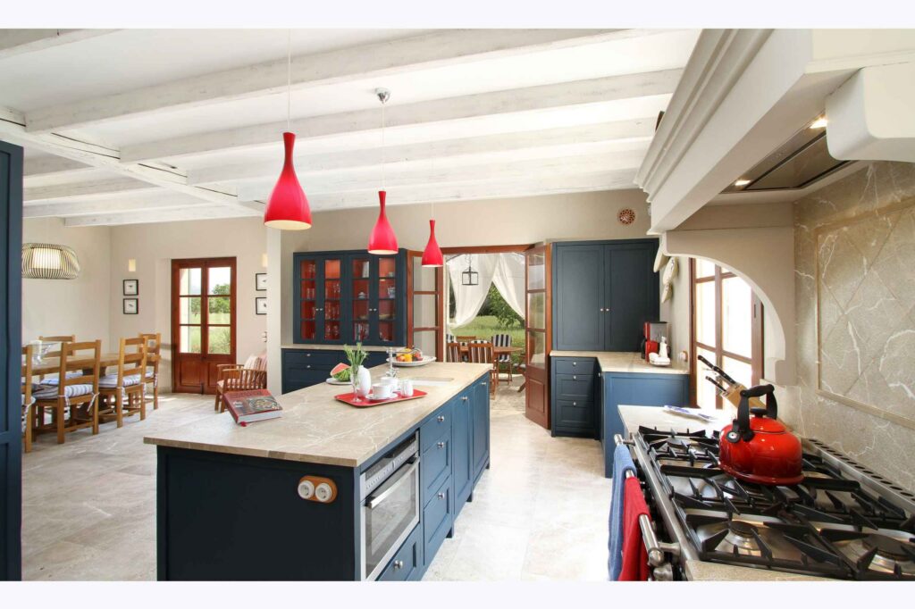 Cocina amplia con isla central, armarios azul marino, lámparas colgantes rojas, fogones y comedor al fondo