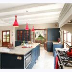 Cocina amplia con isla central, armarios azul marino, lámparas colgantes rojas, fogones y comedor al fondo