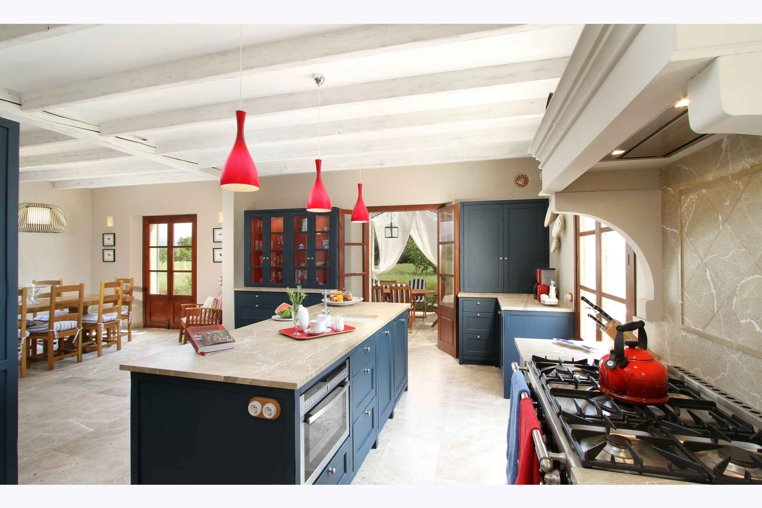 Cocina amplia con isla central, armarios azul marino, lámparas colgantes rojas, fogones y comedor al fondo