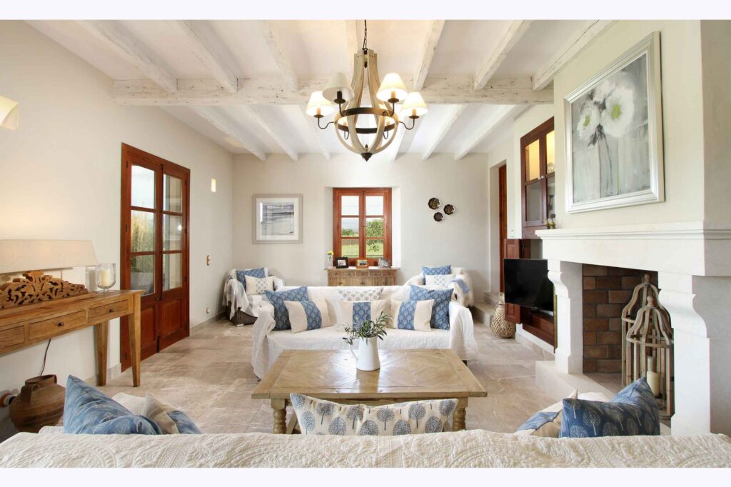 Salón visto desde el sofá, con ventana de madera al fondo, cojines azules y chimenea blanca