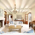 Salón de Can Xandot con vigas blancas, chimenea de piedra clara y sofás blancos con cojines azules