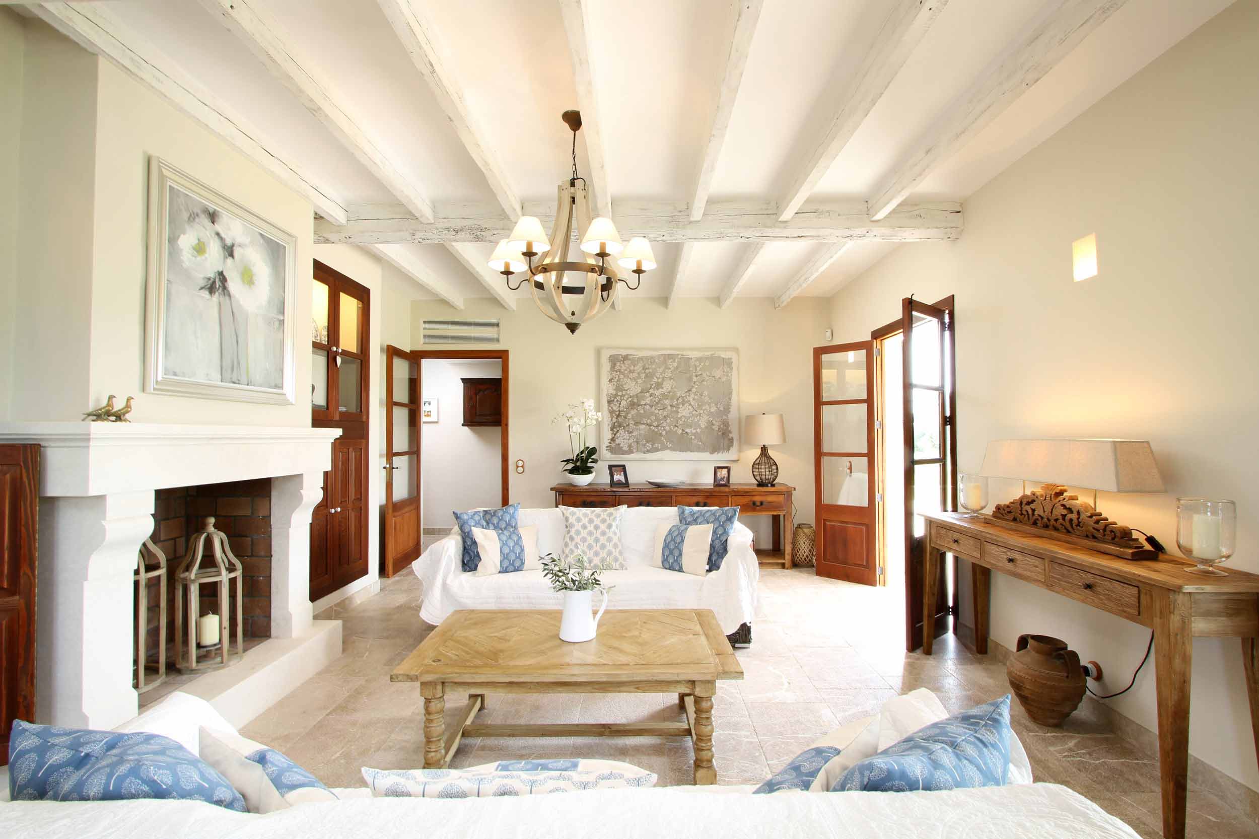 Salón de Can Xandot con vigas blancas, chimenea de piedra clara y sofás blancos con cojines azules