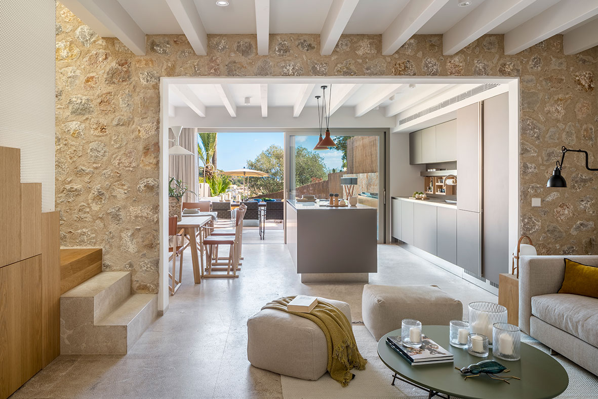 Offener Wohnraum mit Natursteinwand, weißen Deckenbalken und zur Terrasse offener Küche