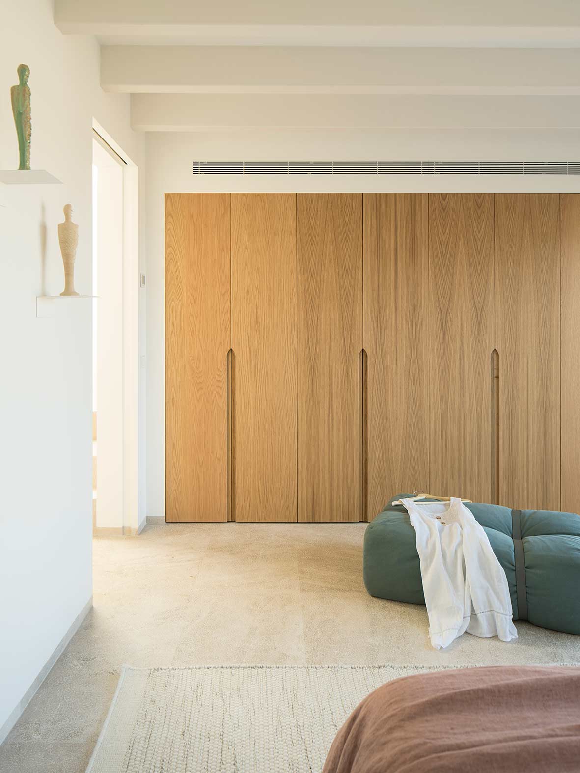 Einbauschrank aus hellem Holz mit integrierten Griffen, grünem Pouf und Teppich in einem hellen Schlafzimmer