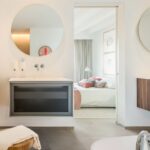 Wandhängendes Waschbecken mit rundem Spiegel im Bad, dahinter Schlafzimmer mit weißem Bett und rosa Kissen