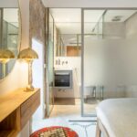 Schlafzimmer mit Glastrennwand zum Bad, Natursteinwand, Schminktisch aus Holz und rotem gemustertem Pouf
