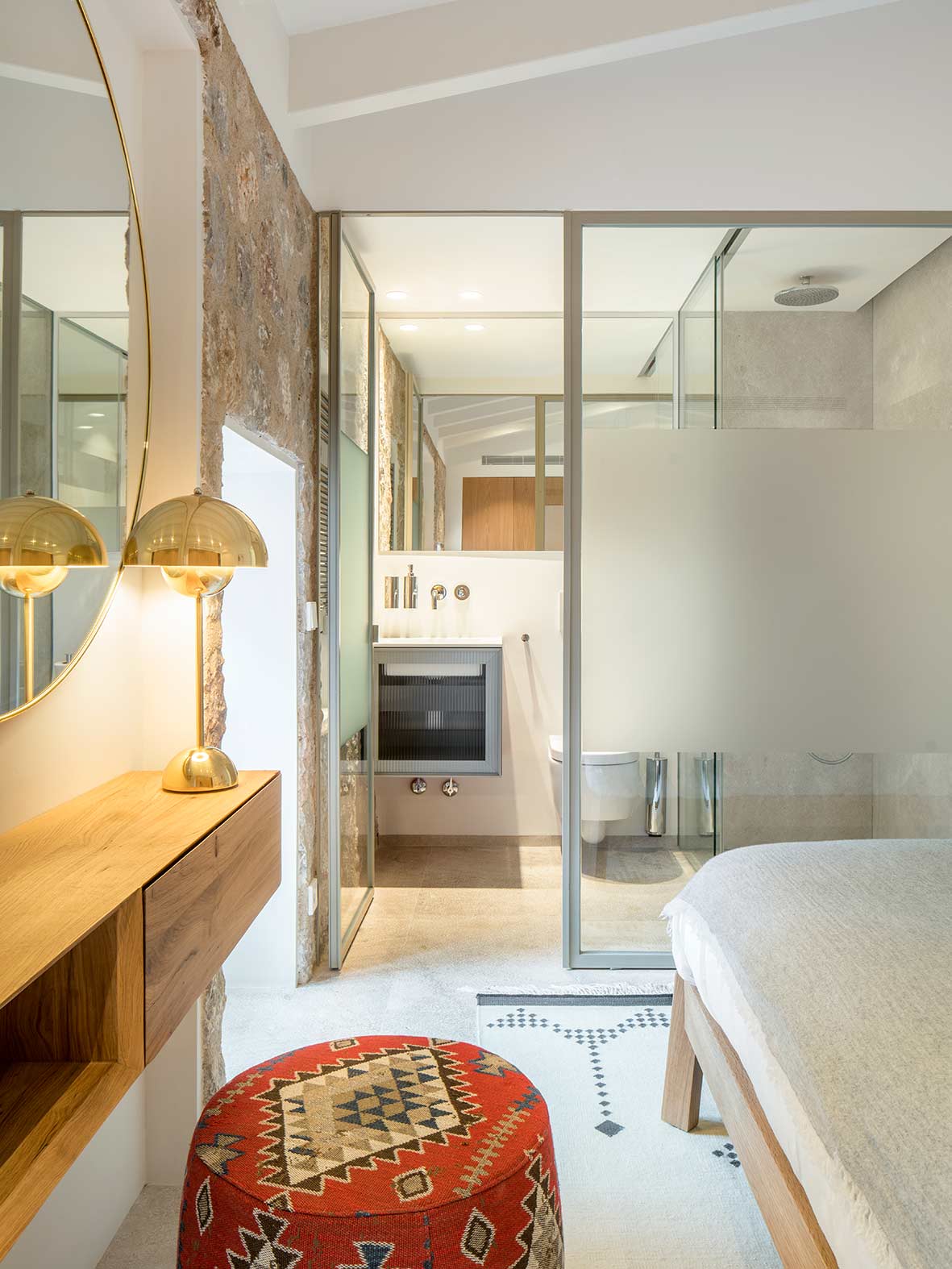Schlafzimmer mit Glastrennwand zum Bad, Natursteinwand, Schminktisch aus Holz und rotem gemustertem Pouf