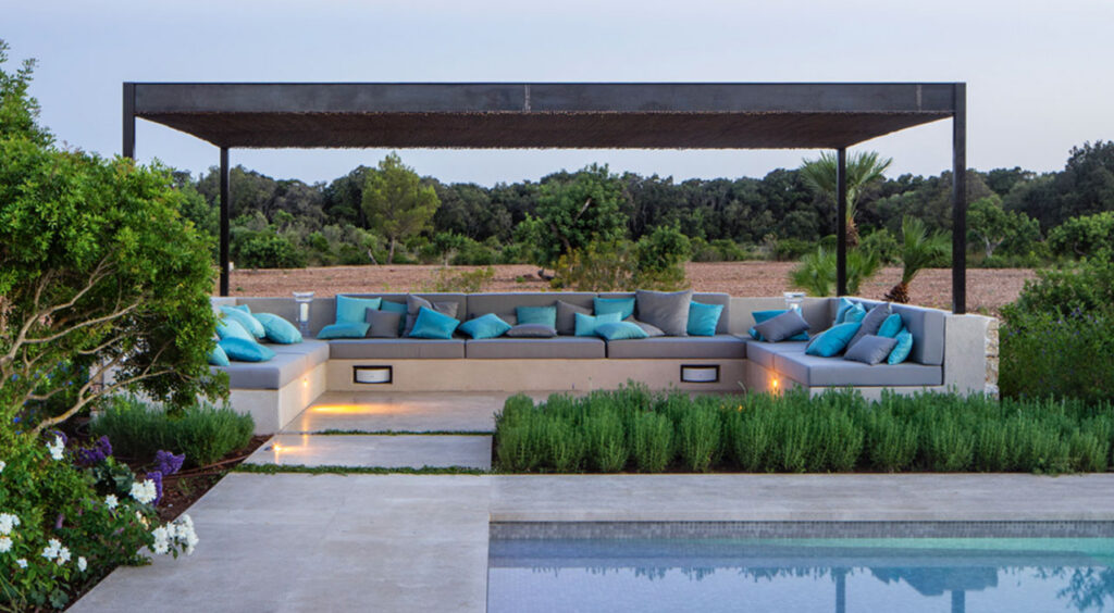 Zona de estar exterior con sofá corrido de obra, cojines turquesa y pérgola metálica junto a la piscina