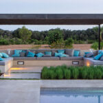 Zona de estar exterior con sofá corrido de obra, cojines turquesa y pérgola metálica junto a la piscina