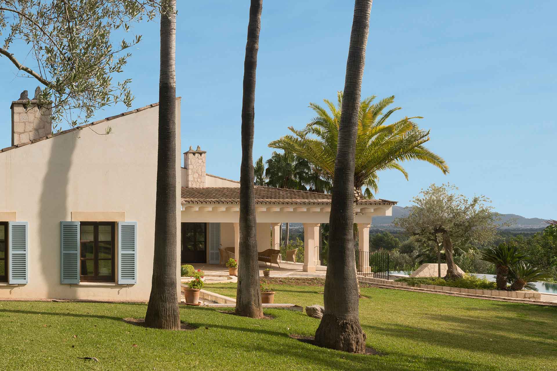 Lateral de la casa con pared clara, persianas azul grisáceo, chimeneas de piedra y porche entre palmeras