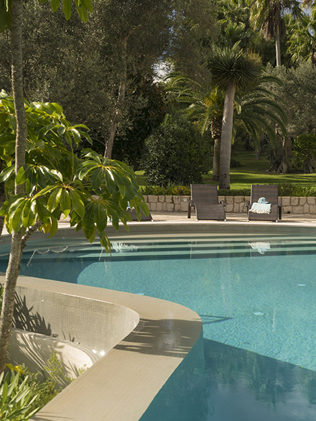 Piscina de agua turquesa con escalones de acceso, tumbonas al fondo, muro de piedra y jardín arbolado