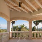 Terraza con arcos y balaustrada, techo abovedado con vigas blancas y vistas a palmeras y montaña