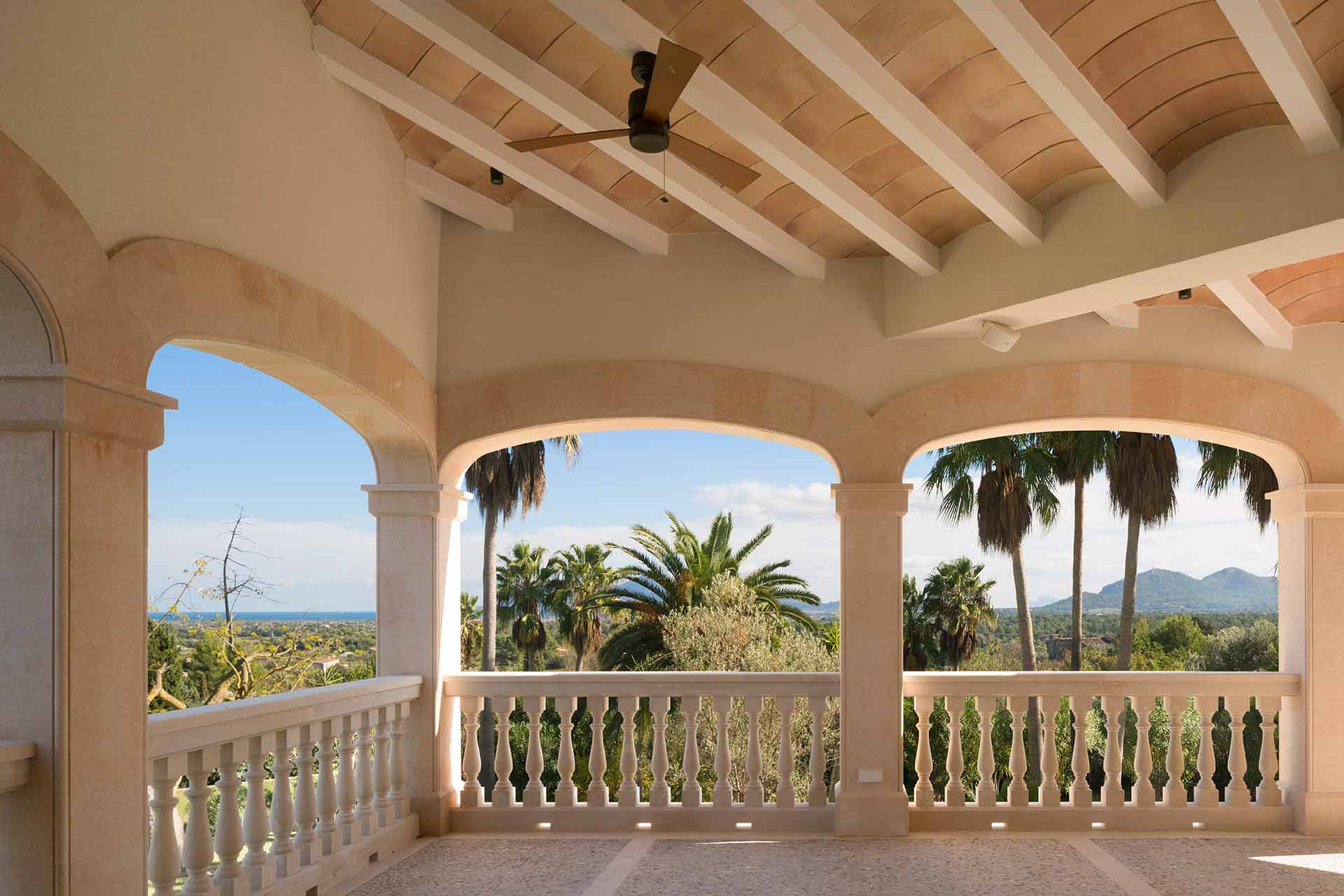 Terraza con arcos y balaustrada, techo abovedado con vigas blancas y vistas a palmeras y montaña
