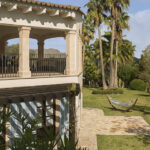 Esquina de la casa con terraza cubierta, pérgola de madera, persianas azules y hamaca sobre el césped