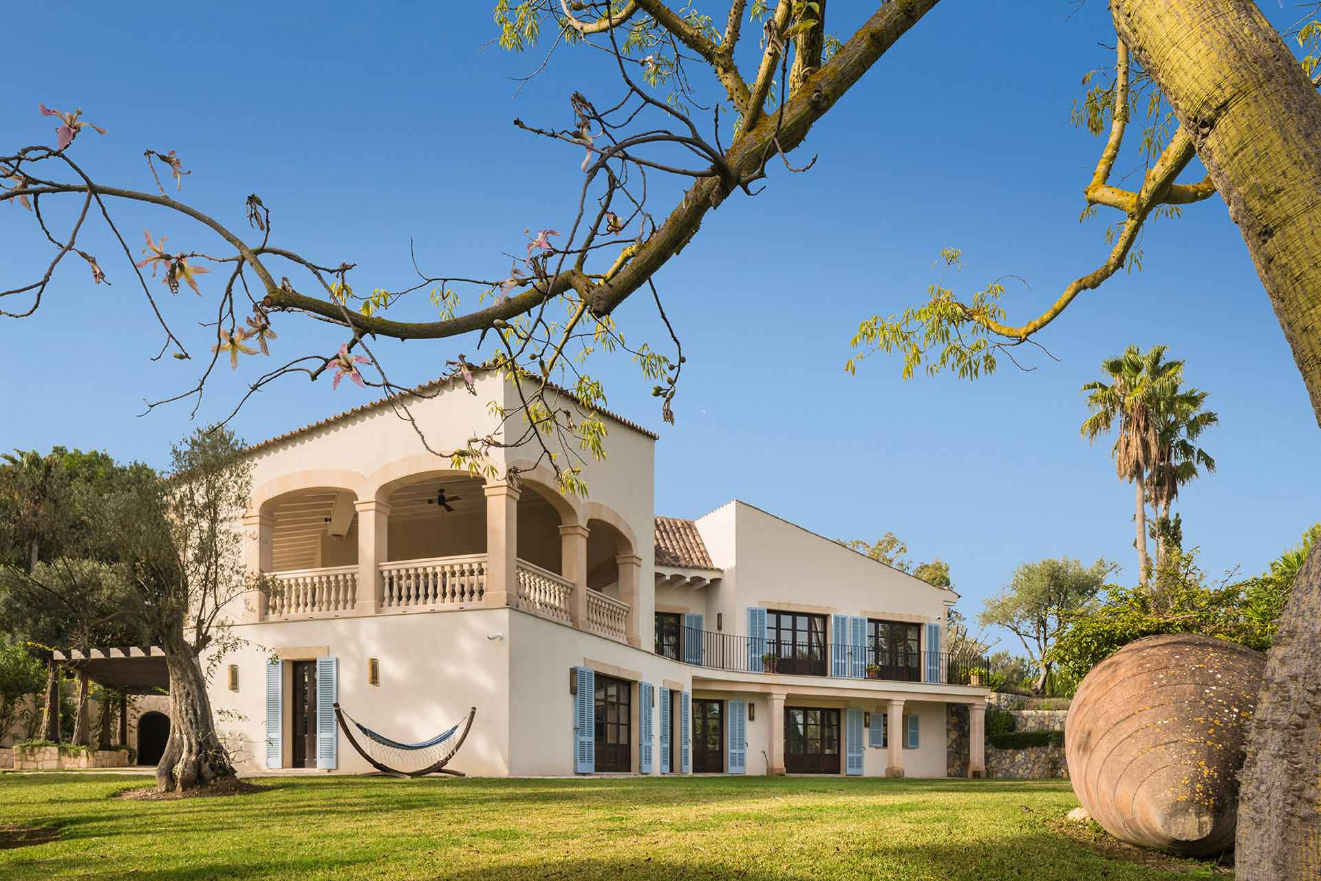 Casa con terraza de arcos y hamaca, con ramas y una gran vasija de barro delante
