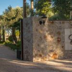 Acceso con muro de piedra, rótulo y farol, y camino de grava entre árboles hacia la casa