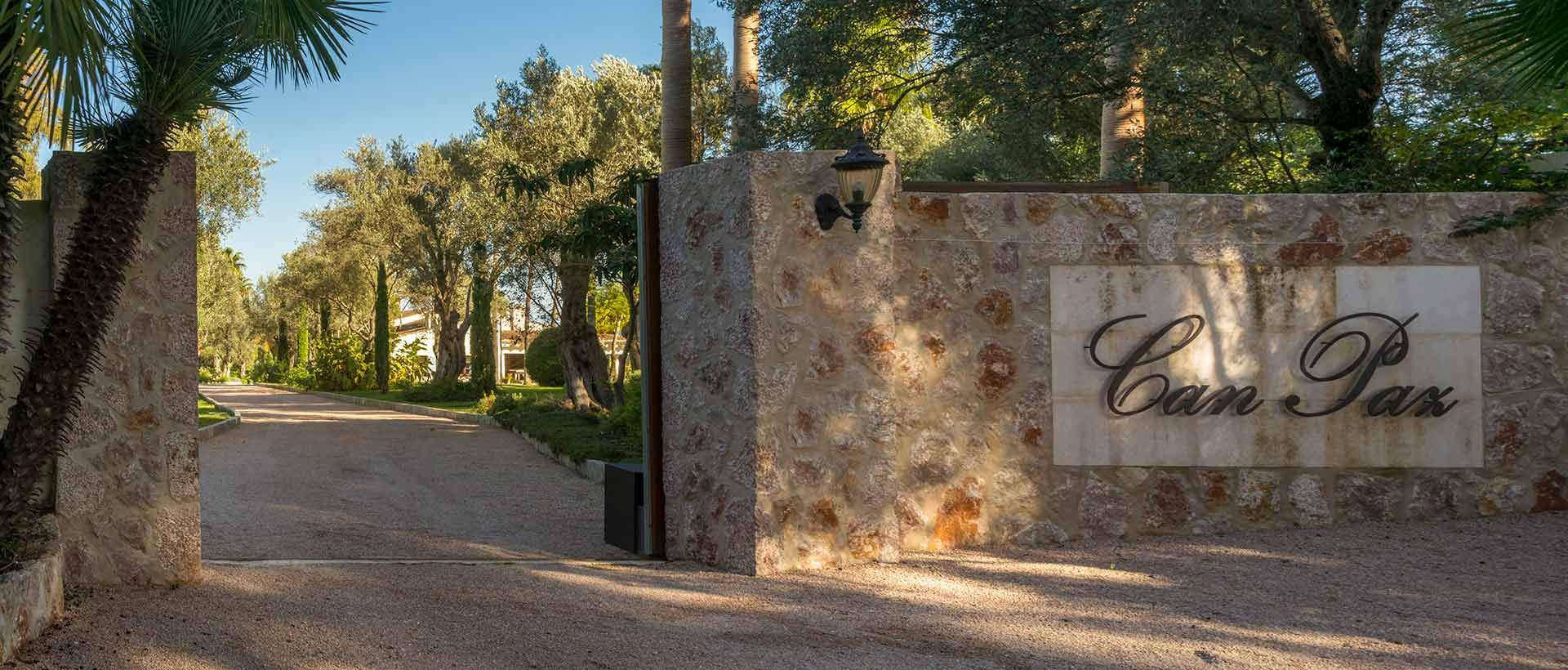 Acceso con muro de piedra, rótulo y farol, y camino de grava entre árboles hacia la casa